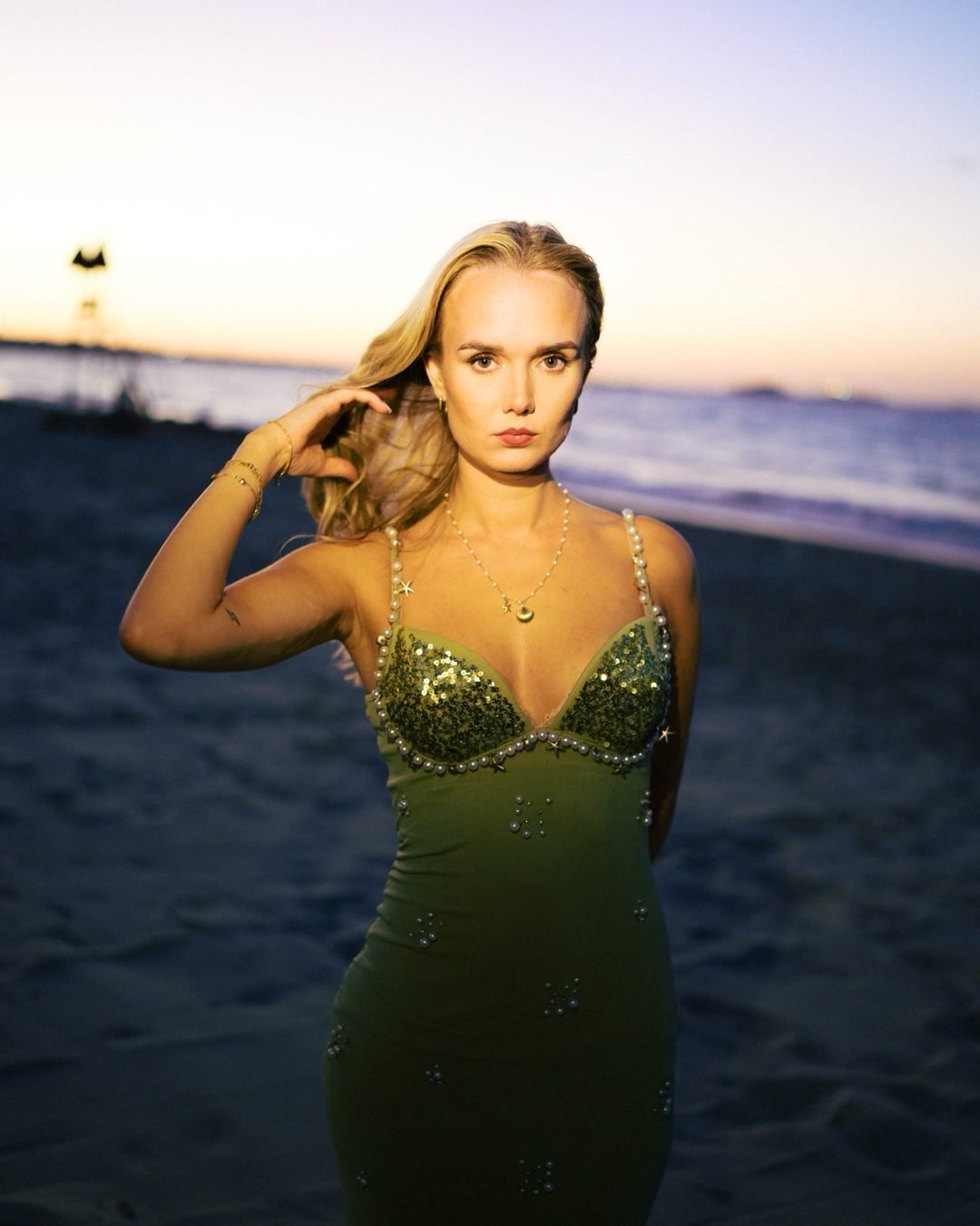 Woman in an olive beaded dress with her hand at her temple against a pastel sunset