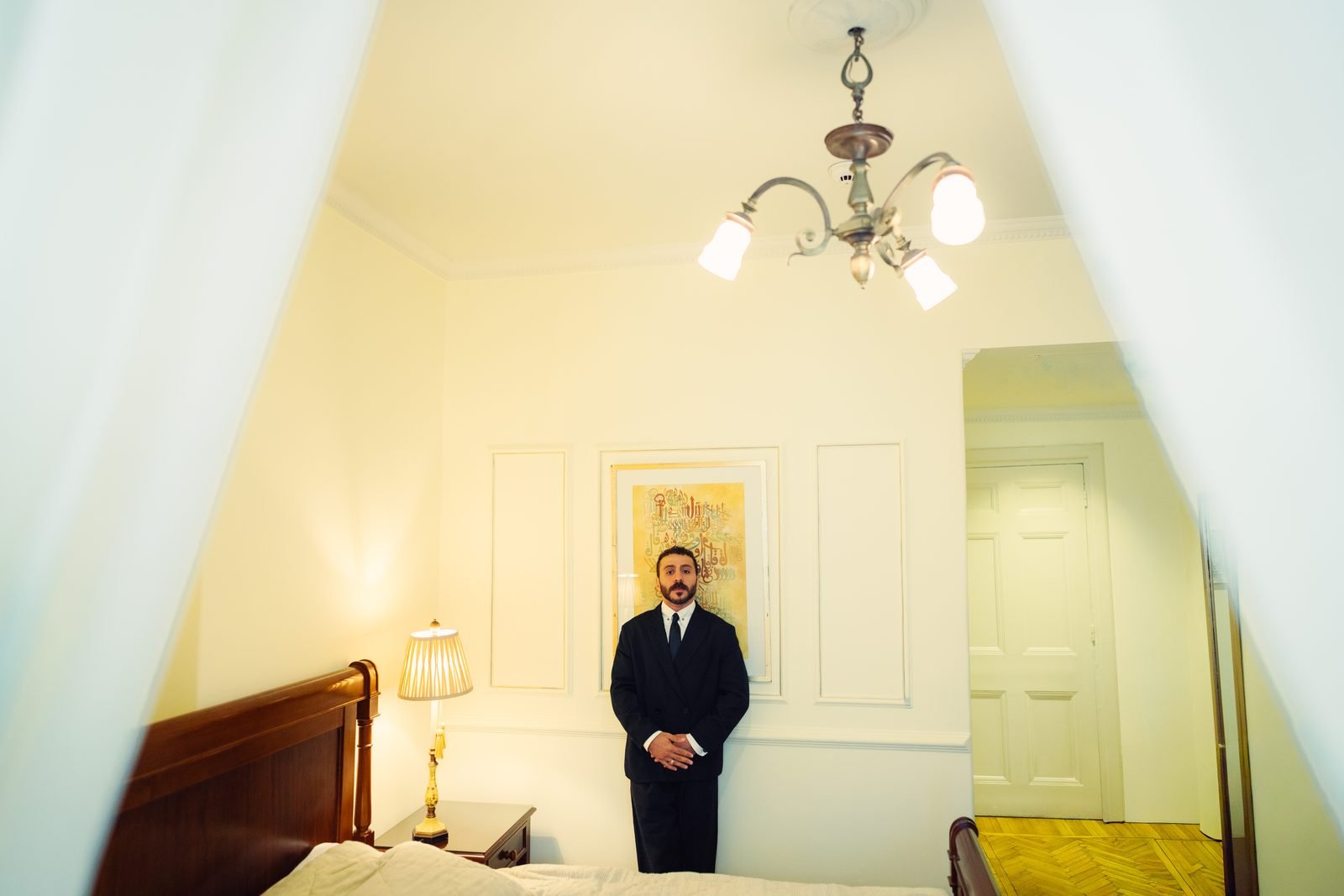 Groom standing in a bedroom framed by curtains