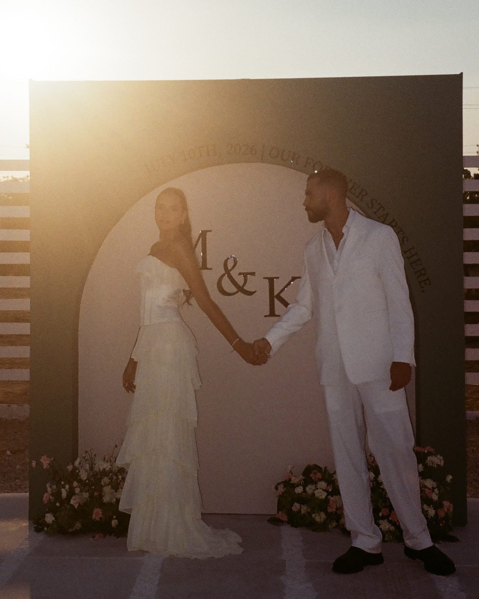Couple holding hands in front of a monogram backdrop at sunset