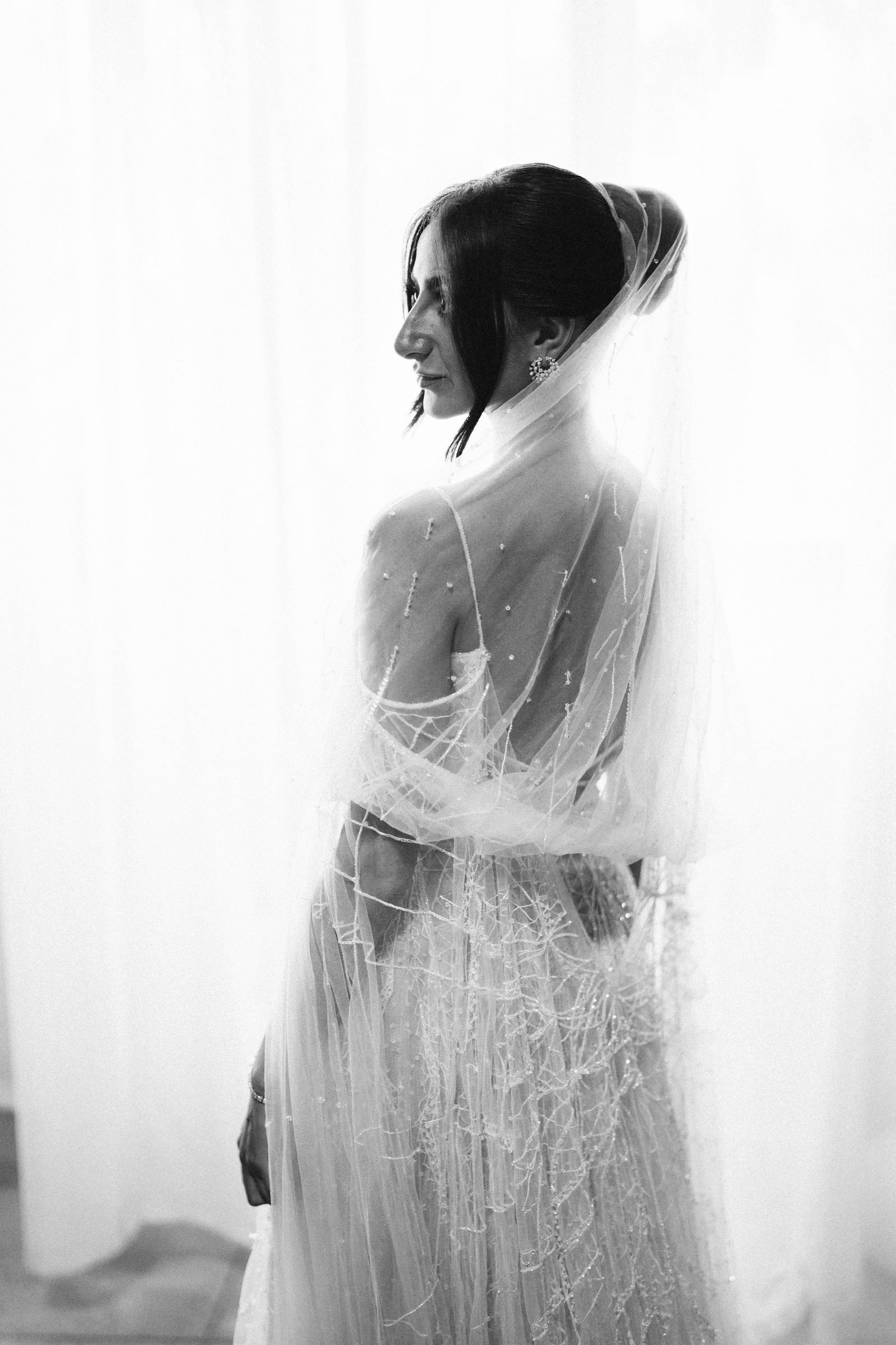 Bride in a beaded veil beside a bright window, black and white