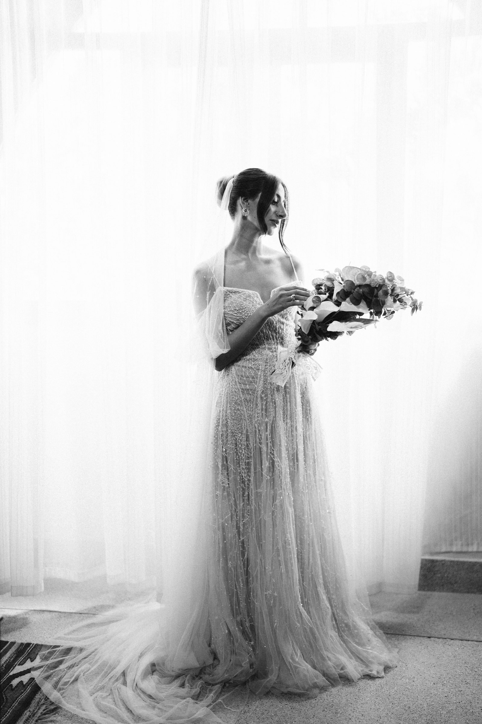 Bride holding a bouquet beside a sunlit window, black and white