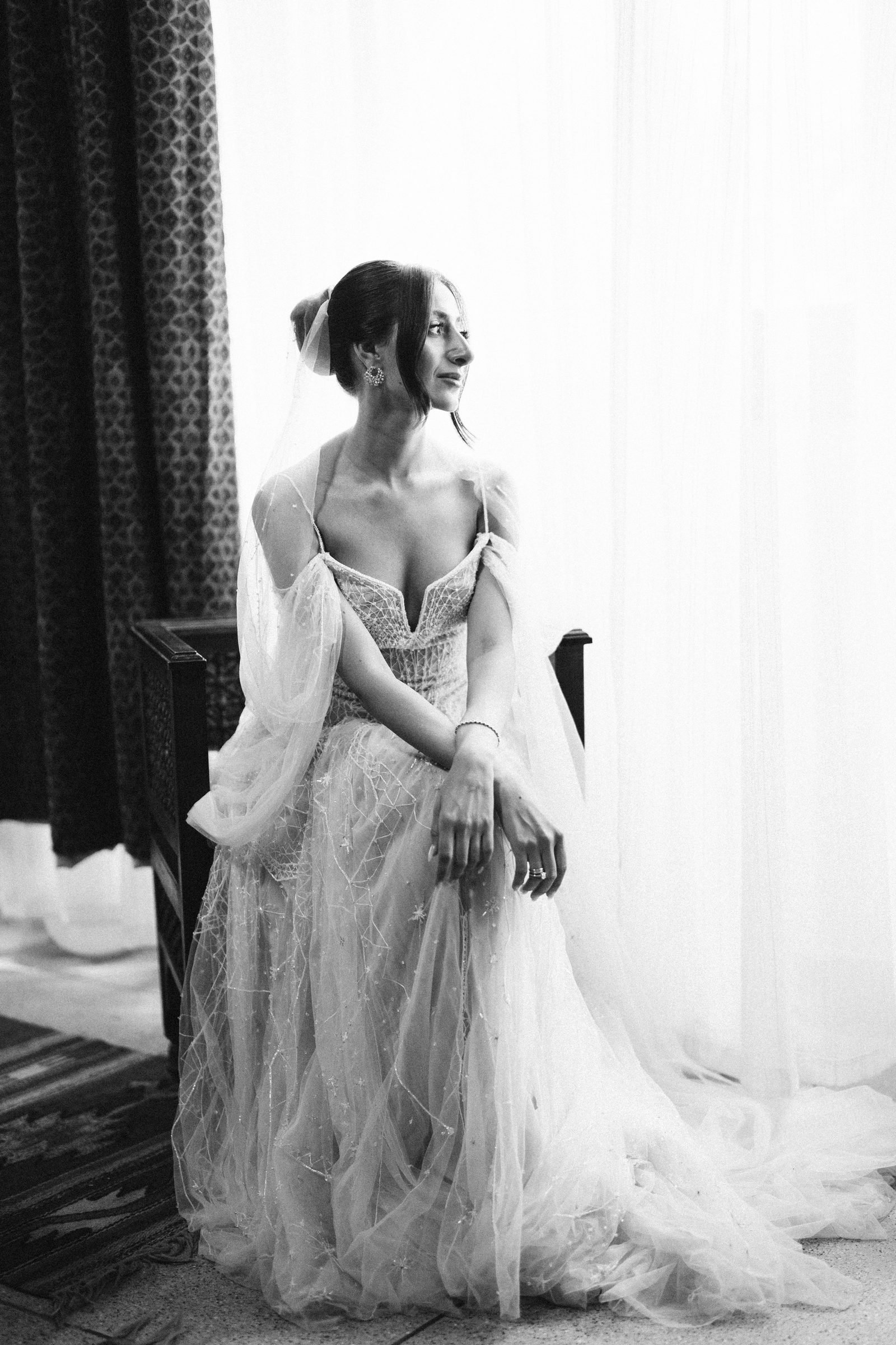 Bride seated in a tulle gown beside sheer curtains, black and white