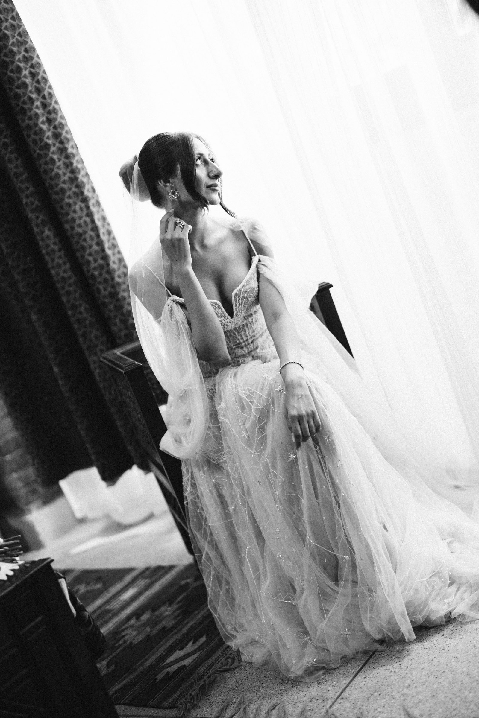 Bride in a tulle gown touching her ear beside sheer curtains, black and white