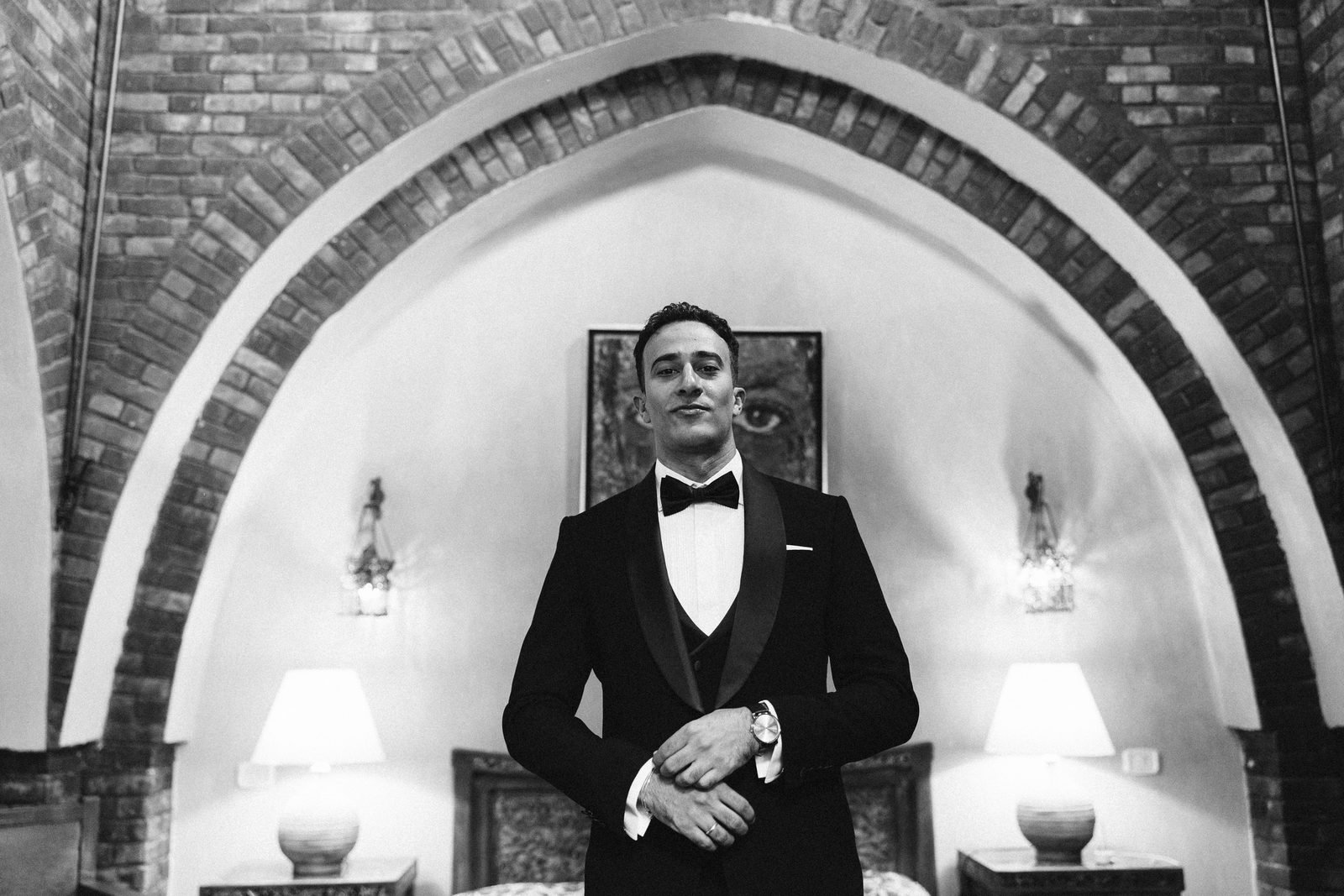 Groom in a bow tie standing beneath a brick arch, black and white